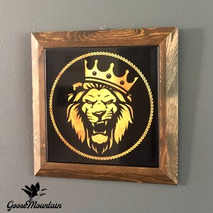 May include: A framed wall art piece with a black background and a gold lion wearing a crown inside a rope circle. The frame is made of dark wood.