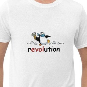 以下が含まれることがあります： 青いスカーフと花を持った漫画のペンギンの白いTシャツ。「revolution」の文字がペンギンのイラストの下に赤で印刷されています。シャツは柔らかい素材で作られています。