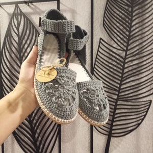 May include: A pair of gray crocheted sandals with a floral design. The sandals have a white sole and a brown tag that says "Yababa Handmade with Love".