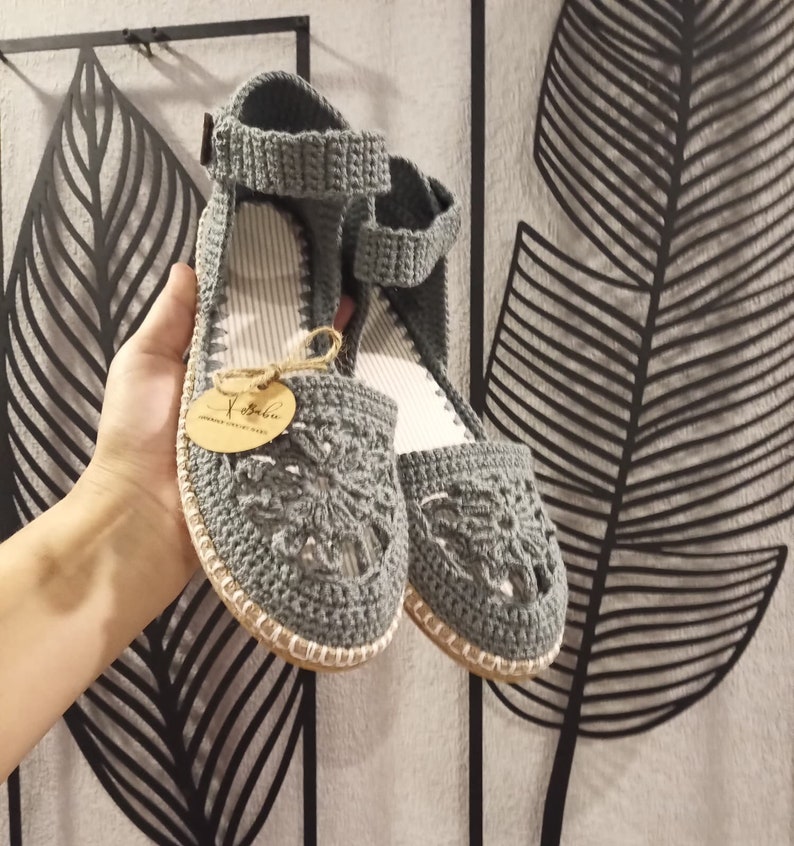 May include: A pair of gray crocheted sandals with a floral design. The sandals have a white sole and a brown tag that says "Yababa Handmade with Love".