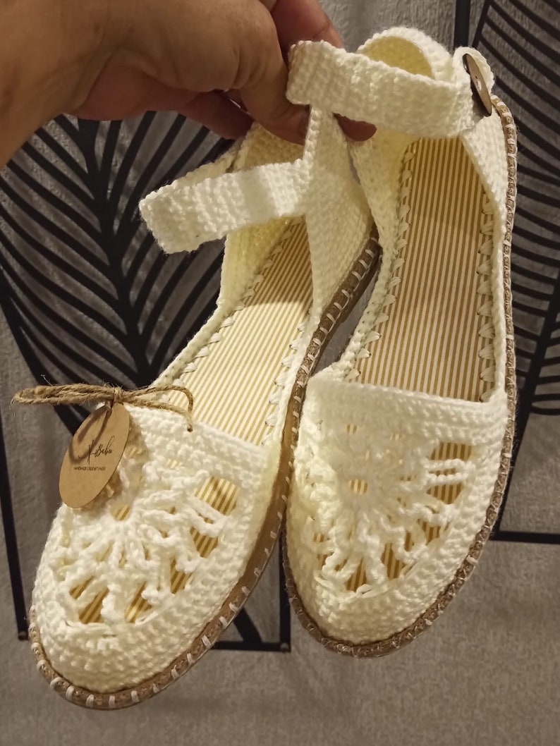 May include: White crocheted sandals with a delicate floral design. The sandals have a brown jute sole and a white strap that goes around the ankle. The sandals are handmade and have a tag that says "Handmade with love".