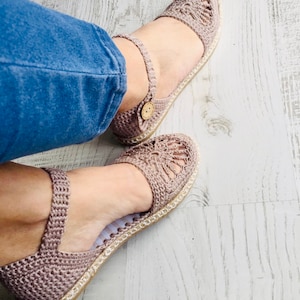 May include: A pair of crocheted sandals with a light brown color. The sandals have a closed toe and a strap around the ankle. The sandals are on a wooden surface.