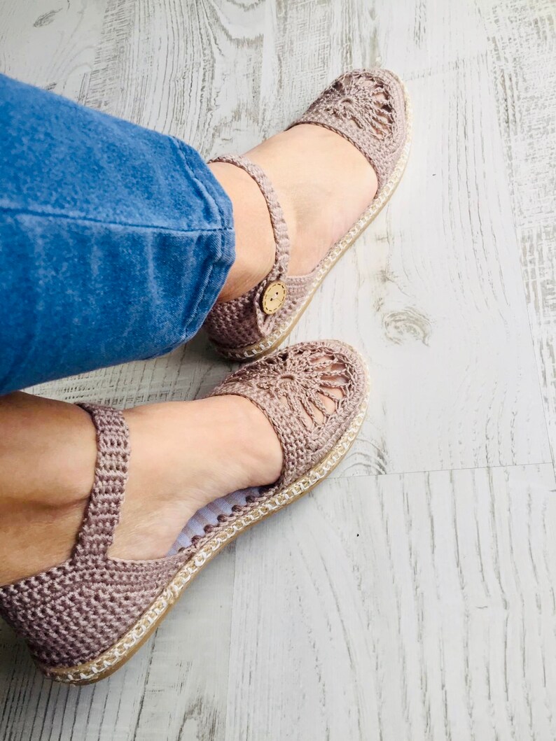 May include: A pair of crocheted sandals with a light brown color. The sandals have a closed toe and a strap around the ankle. The sandals are on a wooden surface.
