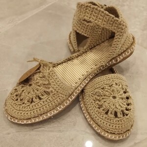 May include: A pair of beige crocheted sandals with a flower design on the toe. The sandals have a white trim around the sole and a strap that goes around the ankle.