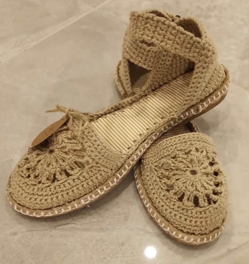 May include: A pair of beige crocheted sandals with a flower design on the toe. The sandals have a white trim around the sole and a strap that goes around the ankle.