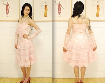 May include: A pink layered lace dress with a satin bodice and spaghetti straps. The dress has a ruffled skirt and is worn with red heels.