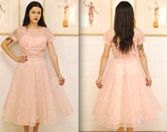 May include: A pink tulle dress with a sheer overlay and a fitted bodice. The dress has a full skirt and short sleeves.