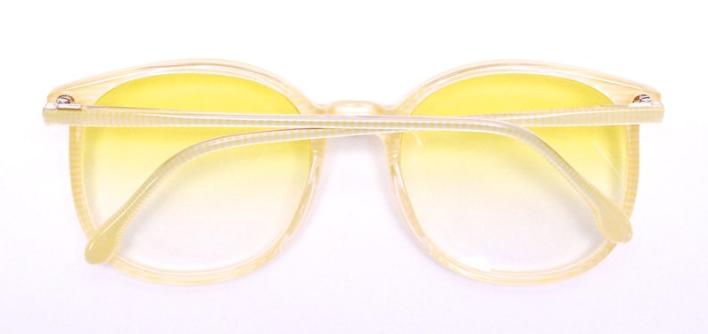 May include: Yellow plastic framed sunglasses with a gradient yellow lens. The sunglasses have a white and yellow striped pattern on the arms.