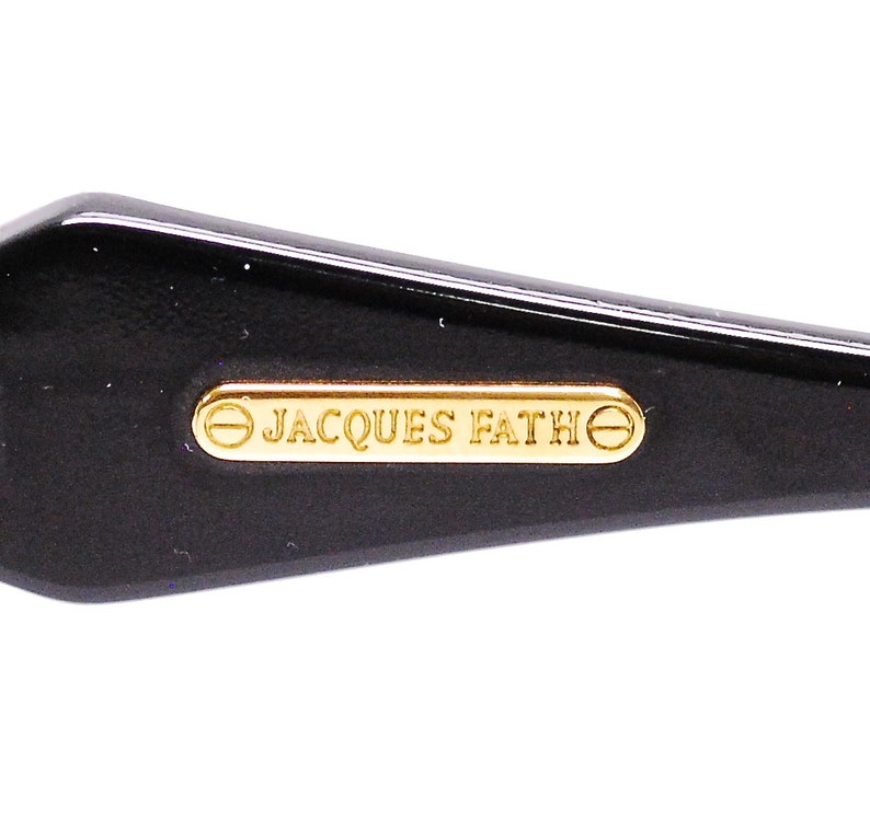 Op de afbeelding: Close-up van een zwart plastic frame met een gouden metalen plaat waarop "JACQUES FATHE" staat.