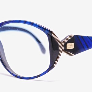Op de afbeelding: Blauwe en zwarte gepatroneerde bril met een gouden accent aan de zijkant. De glazen hebben een blauwe tint.