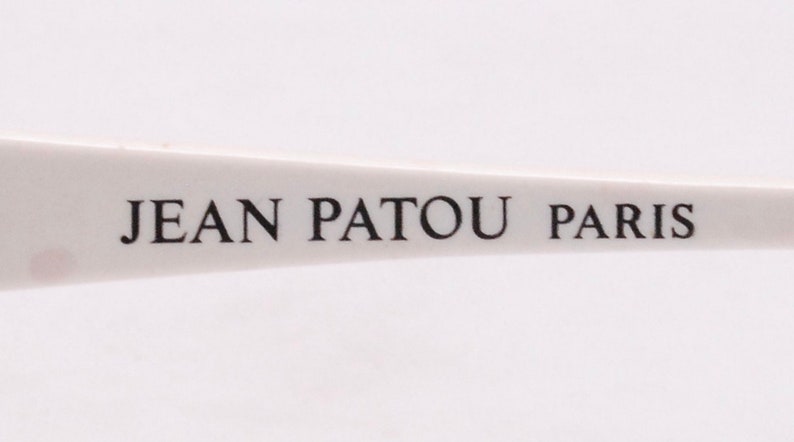 Peut inclure: Gros plan sur une monture en plastique blanche avec le texte "JEAN PATOU PARIS" imprim&eacute; en noir.