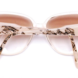 May include: A pair of white plastic sunglasses with a brown lens and a black and white patterned design. The sunglasses have a gold logo on each arm.