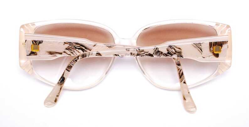 May include: A pair of white plastic sunglasses with a brown lens and a black and white patterned design. The sunglasses have a gold logo on each arm.
