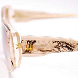 May include: Beige and brown patterned sunglasses with a gold metal accent on the side. The sunglasses have a rectangular shape and a thin frame.