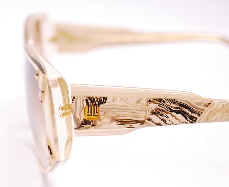 May include: Beige and brown patterned sunglasses with a gold metal accent on the side. The sunglasses have a rectangular shape and a thin frame.