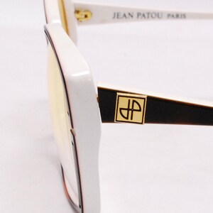 Peut inclure: Lunettes de soleil blanches avec un logo Jean Patou Paris noir et or sur la branche. Les lunettes ont une monture carr&eacute;e et des verres jaunes.