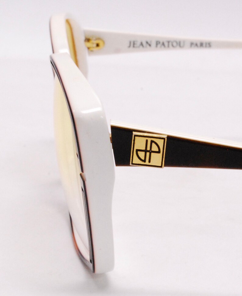 Peut inclure: Lunettes de soleil blanches avec un logo Jean Patou Paris noir et or sur la branche. Les lunettes ont une monture carr&eacute;e et des verres jaunes.