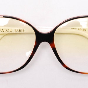 Peut inclure: Lunettes de vue en &eacute;caille brune et blanche avec un pont et des branches blancs. Les montures pr&eacute;sentent un motif g&eacute;om&eacute;trique noir et blanc. Les verres sont teint&eacute;s en jaune. Le texte "JEAN PATOU PARIS" est imprim&eacute; sur la monture gauche. Le texte "P/F SP 39 COL 531" est imprim&eacute; sur la monture droite.