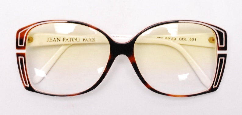 Peut inclure: Lunettes de vue en &eacute;caille brune et blanche avec un pont et des branches blancs. Les montures pr&eacute;sentent un motif g&eacute;om&eacute;trique noir et blanc. Les verres sont teint&eacute;s en jaune. Le texte "JEAN PATOU PARIS" est imprim&eacute; sur la monture gauche. Le texte "P/F SP 39 COL 531" est imprim&eacute; sur la monture droite.