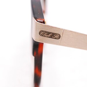 Op de afbeelding: Close-up van een schildpadframe met een zilveren metalen arm. De arm heeft de tekst "Club R" gegraveerd.
