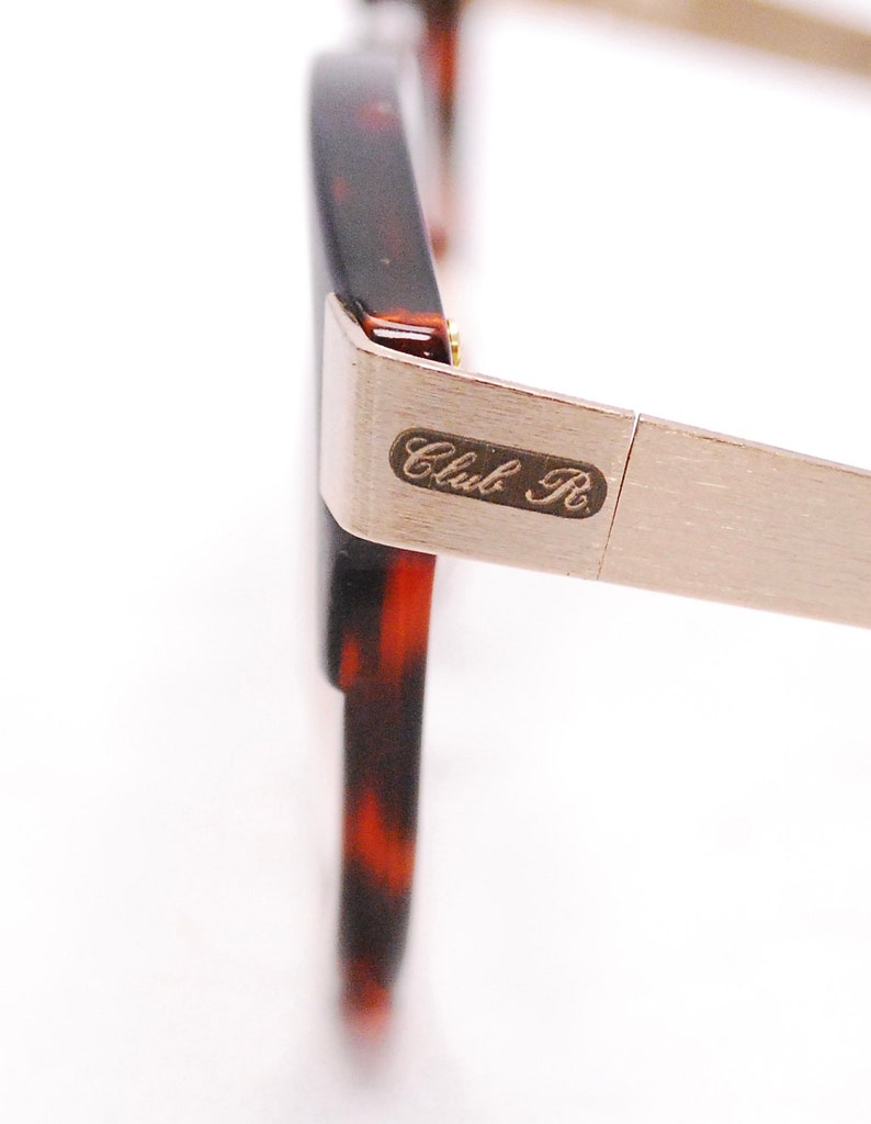 Op de afbeelding: Close-up van een schildpadframe met een zilveren metalen arm. De arm heeft de tekst "Club R" gegraveerd.