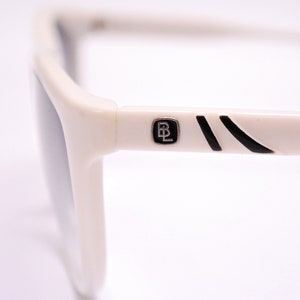 May include: White sunglasses with black accents and a black and silver logo that says "BL" on the side.