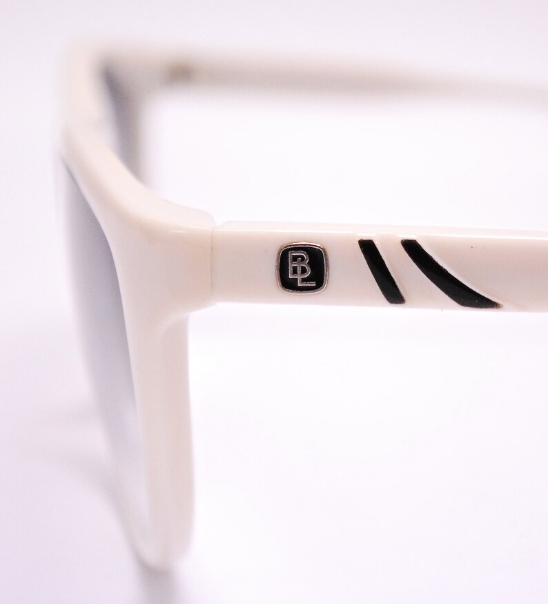 May include: White sunglasses with black accents and a black and silver logo that says "BL" on the side.