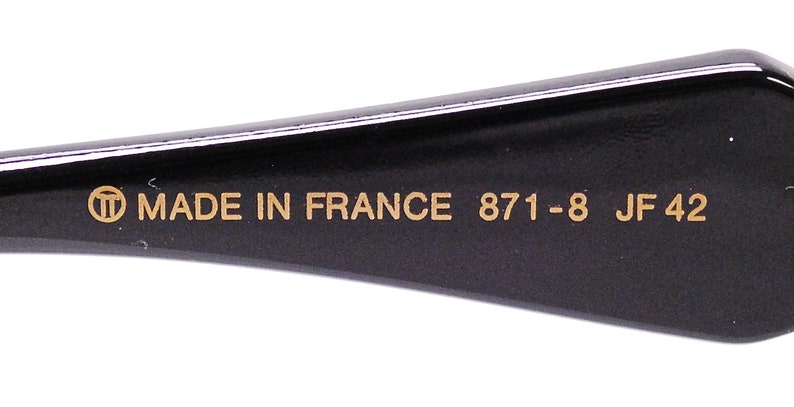 Op de afbeelding: Close-up van de zwarte poot van een bril met gouden letters die "MADE IN FRANCE 871 -8 JF 42" lezen.