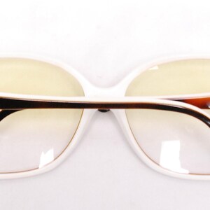 Peut inclure: Lunettes rectangulaires &agrave; monture blanche avec des branches en &eacute;caille de tortue brune et des accents dor&eacute;s. Le nom de la marque est visible sur les branches.