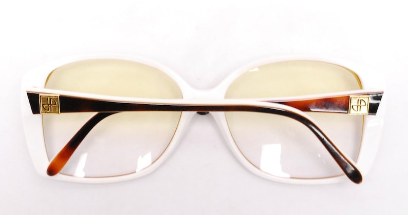 Peut inclure: Lunettes rectangulaires &agrave; monture blanche avec des branches en &eacute;caille de tortue brune et des accents dor&eacute;s. Le nom de la marque est visible sur les branches.
