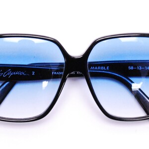 May include: Black square-shaped sunglasses with blue lenses and a marble pattern on the frames. The sunglasses have a signature "Cavolo Capucci" inscription on the left lens and "MARBLE 58-13-140mm" on the right lens.