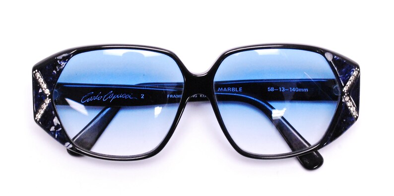 May include: Black square-shaped sunglasses with blue lenses and a marble pattern on the frames. The sunglasses have a signature "Cavolo Capucci" inscription on the left lens and "MARBLE 58-13-140mm" on the right lens.