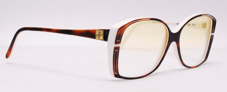 Peut inclure: Lunettes de vue blanches et &eacute;caille de tortue avec un accent dor&eacute; sur la branche. Les verres sont clairs et la monture a une forme &eacute;paisse et arrondie.