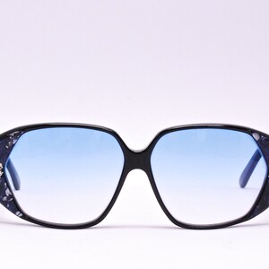 May include: Black sunglasses with blue lenses and a decorative pattern on the frames. The pattern includes small, clear rhinestones.