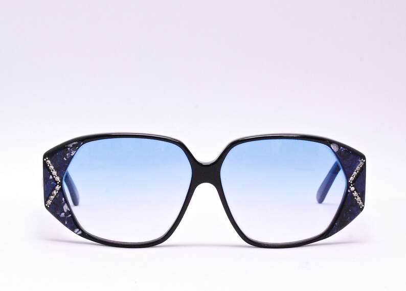 May include: Black sunglasses with blue lenses and a decorative pattern on the frames. The pattern includes small, clear rhinestones.