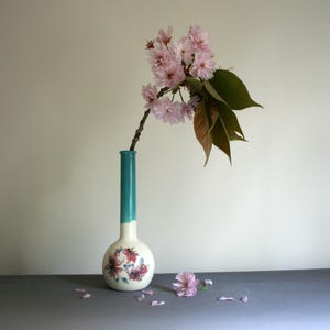 May include: A tall, slender ceramic vase with a round base and a long, narrow neck. The neck is glazed in a solid teal colour, while the base is off-white with a floral pattern in shades of pink and blue. A branch with pink blossoms and green leaves.