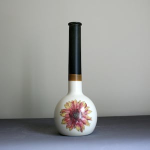 May include: A ceramic vase with a round base and a tall, slender neck. The base is white with a large, detailed sunflower illustration in shades of yellow, orange, and red. The neck is matte black with a thin brown band near the base of the neck.
