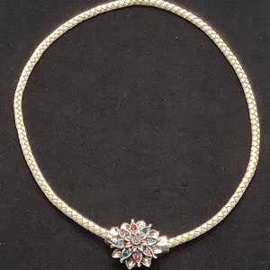 May include: A white leather choker necklace with a silver flower charm. The flower charm has red, blue, and clear rhinestones.