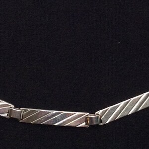 May include: A silver bracelet with rectangular links, each with diagonal etched lines. The bracelet has a clasp at each end. The design is simple and elegant. The bracelet's length is approximately 20 cm.