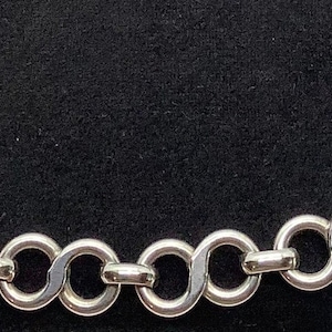 May include: A silver bracelet featuring a chain of interlocking infinity symbol links. The bracelet has a lobster clasp. Each link is about 2 cm in diameter. The bracelet is shown against a black background.