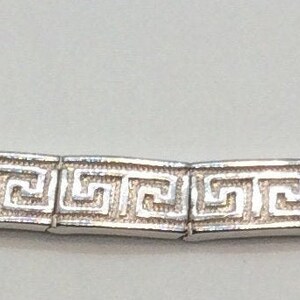 May include: Silver bracelet with a repeating Greek key pattern. The bracelet is made up of rectangular links, each showing the classic meander design. A clasp fastens the bracelet. This jewellery item is suitable for various occasions.