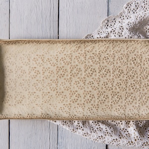 Puede incluir: Una fuente rectangular de cerámica crema con una superficie texturizada y un patrón punteado. La fuente tiene un borde ligeramente irregular y está sobre un paño de encaje blanco.