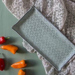 May include: A rectangular, light blue ceramic serving platter with a speckled pattern and a dark blue rim. The platter is displayed on a lace cloth with several red and orange mini bell peppers.
