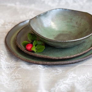 May include: A set of three green ceramic plates with a rustic finish. The plates are stacked on top of each other, with a small sprig of greenery and a red berry resting on the top plate.