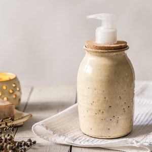 Könnte beinhalten: Ein Keramik-Seifenspender mit einem Korkverschluss und einer Pumpe. Der Spender ist mit Braun und Weiß gesprenkelt und hat einen rustikalen Look. Er steht auf einem weißen Handtuch.