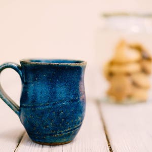 May include: A blue ceramic mug with a speckled glaze and a rounded shape. The mug has a handle and is sitting on a white wooden surface.