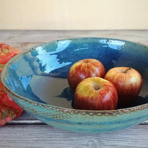 May include: A large, blue ceramic bowl with a textured rim, filled with three red apples. The bowl is sitting on a wooden surface.