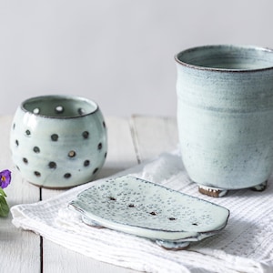 Puede incluir: Un juego de baño de cerámica azul claro. El juego incluye un vaso, un jabonera y un pequeño portavelas redondo con puntos oscuros. Los artículos están sobre una toalla blanca, con una ramita de flores moradas.