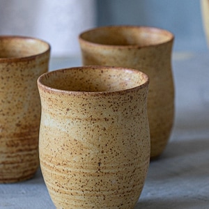 May include: Three ceramic mugs with a speckled brown and yellow glaze. The mugs are all slightly different shapes and sizes.