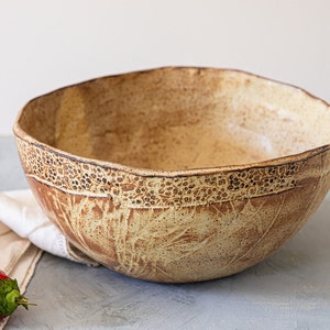 May include: A large, handmade ceramic bowl with a light brown glaze and a decorative pattern around the rim. The bowl is slightly irregular in shape and has a rustic, handmade look.